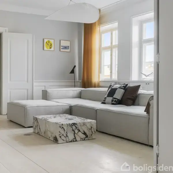 En grå hjørnesofa står i en lys stue med hvide vægge og gul gardin. En marmorbord står foran, og to små billeder hænger på væggen. En lampe er tændt.