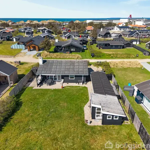 Et fritliggende hus med have ses fra oven, omgivet af andre lignende huse. Baggrunden viser en blå himmel og havet i horisonten.