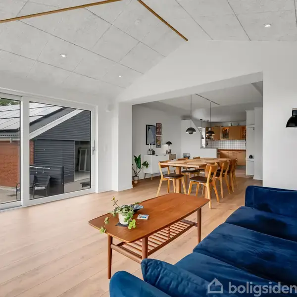 En stue med en blå sofa og et træbord, der vender mod store vinduer. Stuen er forbundet med en spisestue med et spisebord og stole. Udsigt til en terrasse og husfacade udenfor.