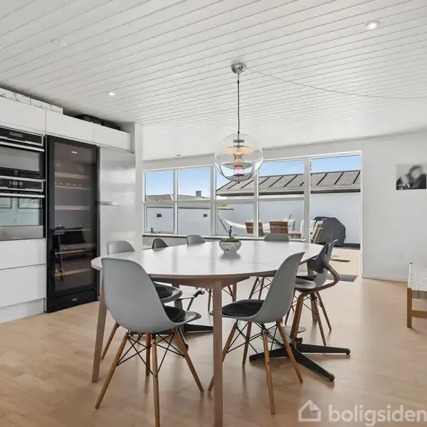 En rund spisebord med sorte stole står i et lyst, moderne køkken. Store vinduer giver udsigt til en terrasse. En hængende lampe og kunst pryder rummet.