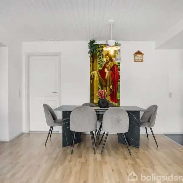Spisebord med seks polstrede stole står på et trægulv. Over bordet hænger en lampe, og på væggen ses et farverigt billede af en religiøs figur. Værelset er enkelt indrettet.