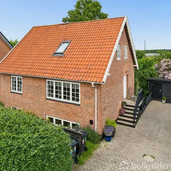 Rød murstenshus med rødt sadeltag, vinduer og dør. Det står på flisebelagt indkørsel omgivet af hække og planter. Der ses en sort trappe ved siden af hoveddøren.