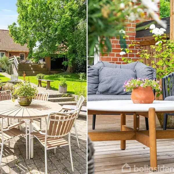 Rundt havebord med stole står på en terrasse i solskinsvejr. Til højre er der en sofa med puder og et bord med blomster, omkranset af grønne planter og træer.