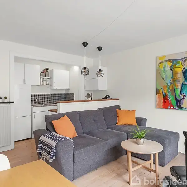 Grå sofa med orange puder står i en stue ved siden af et rundt bord med en plante. Farverigt maleri hænger på væggen. Moderne åbent køkken i baggrunden.