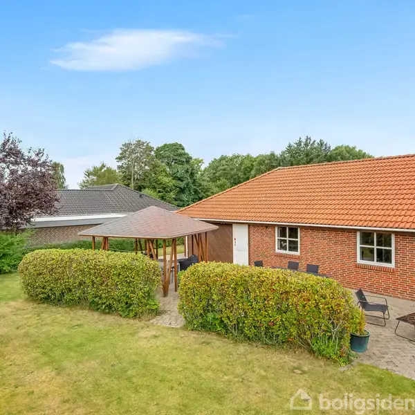 Rød murstenshus med rød tagstenstag omgivet af en have. Havemøbler står på en fliseterrasse foran huset. En pavillon med hegn af buske ses i baggrunden under en blå himmel.