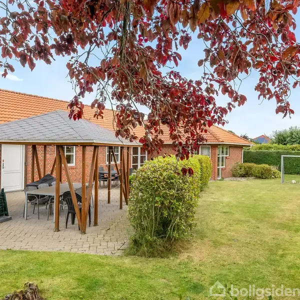 Pavillon står midt i en have, omgivet af grønne buske og træer med røde blade. Bord med stole er placeret under pavillonen. I baggrunden er et fodboldmål.