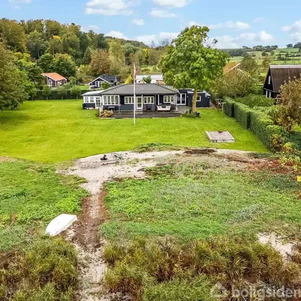 En sort-hvid bygning omgivet af en velholdt græsplæne med træer, placeret i en landskabelig, grøn dal med flere huse i nærheden. En jordsti fører gennem området.