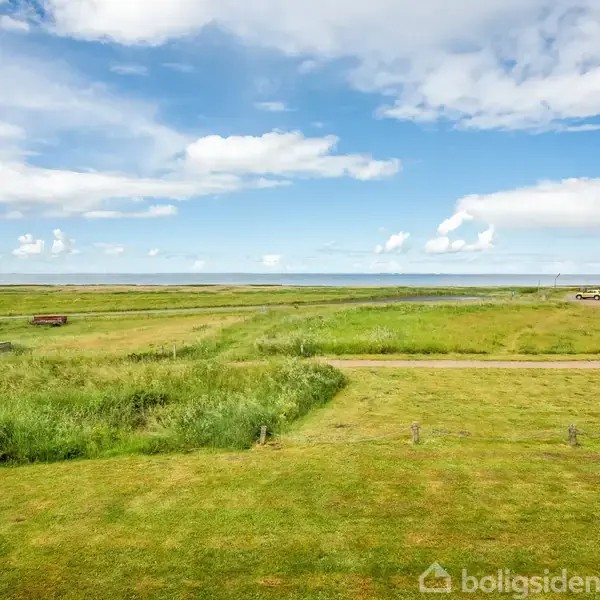 Grønne marker strækker sig mod en horisontlinje med havudsigt, omgivet af en klar blå himmel med hvide skyer, og en bygning ses til højre.