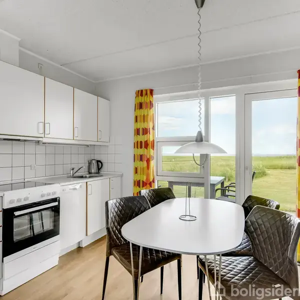 Køkken med hvidt interiør, spisebord og fire stole. Sollys strømmer ind gennem et stort vindue med farverige gardiner. Udsigt til grønt landskab udenfor.