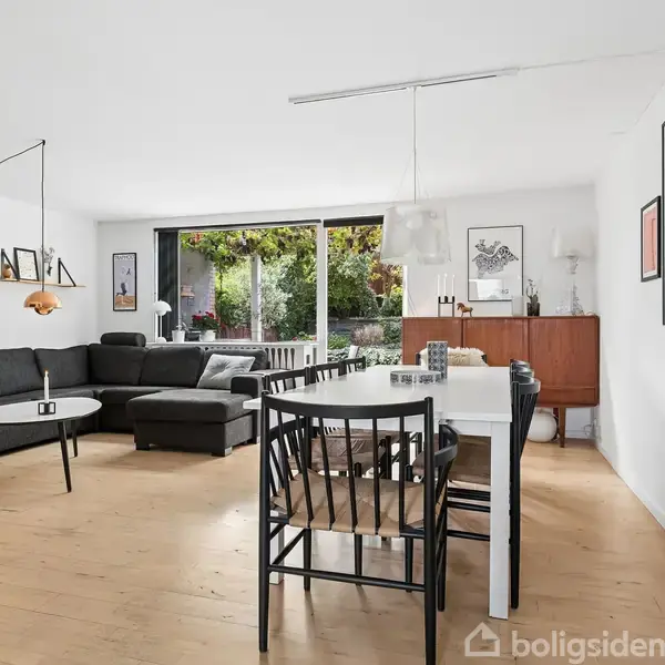 En stue med en sort sofa i hjørnet, et spisebord med stole, og store vinduer med udsigt til en grøn have. Indretningen er moderne med minimalistiske møbler.