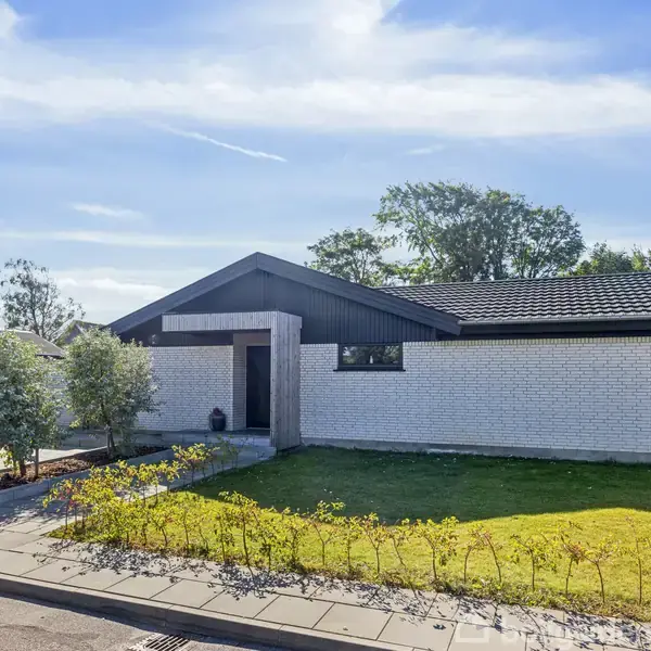 En moderne, hvid murstensvilla står stille omgivet af en velplejet græsplæne og små buske. Beliggende i et stille beboelsesområde med blå himmel og få træer i baggrunden.