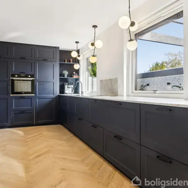 Sort køkken med indbyggede skabe og ovn, skuffer langs væggen, moderne pendellamper, stor vindue med udsigt til udendørsområde, lys trægulv, minimalistisk design, hvid bordplade.