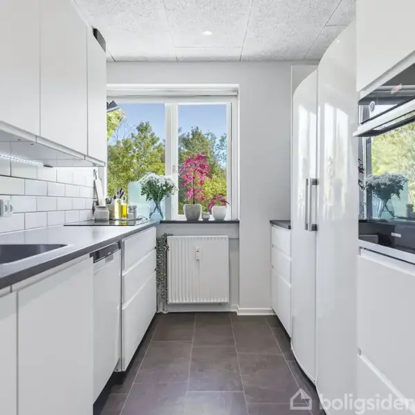 Et moderne køkken med hvide skabe og mørke bordplader, har en stålvask og indbyggede ovne. Sollys strømmer ind gennem et vindue med udsigt til træer og blomster udenfor.