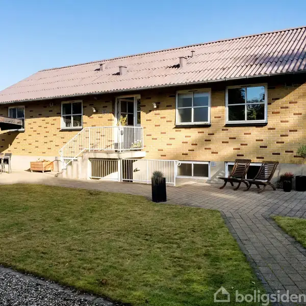 Et murstenshus med rødt tegltag står i en velholdt have. Foran huset er der en terrasse med havemøbler, og stierne er af stenbelægning.