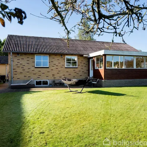 Et hus med murstensfacade står på en grøn græsplæne. Ved huset er der en markise og en udestue af træ med vinduer. Sollys fylder den omkringliggende have.