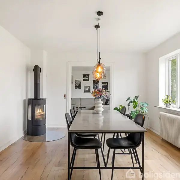 En spisebord i moderne design er placeret i et lyst rum. Over bordet hænger to lamper. I nærheden ses en brændeovn. Vinduet til højre giver udsigt til grønne omgivelser.