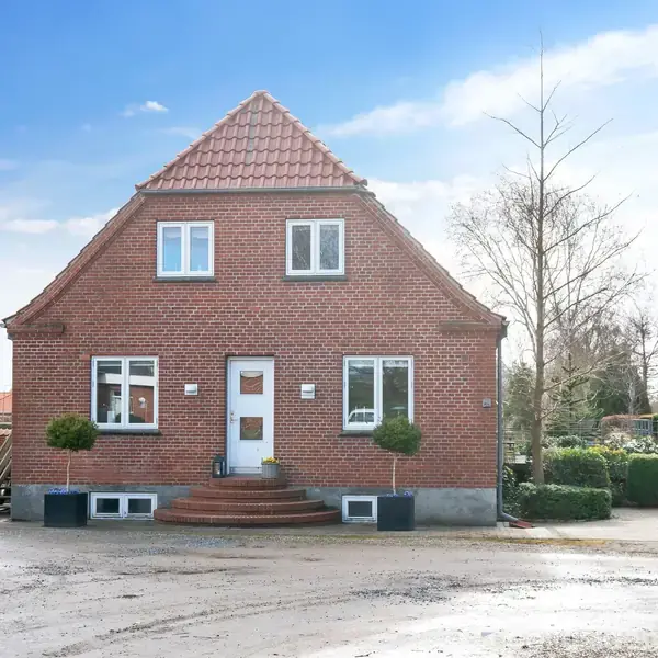 Rødt murstenshus ligger stille ved en grusvej. Huset har en klassisk gavlform og en trappe op til hoveddøren. Omgivet af små buske og vintertræer.