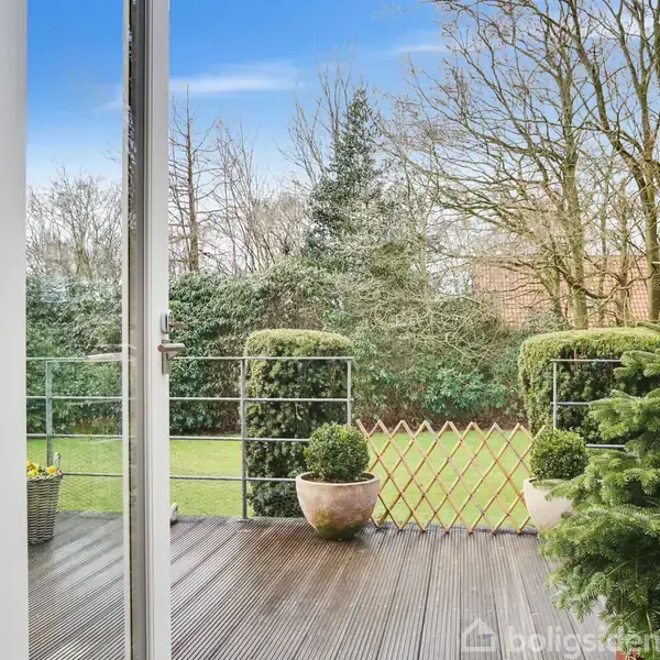 Glasdøre åbner mod en terrasse med plantekasser og grønne buske, omgivet af en velholdt have med græsplæne og træer. Himlen er blå med spredte skyer.