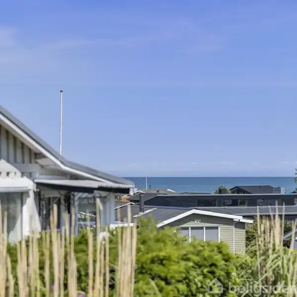 Et gråt hus med skiltet "Fjeldet" i venstre side står stille i et fredeligt boligområde. Havet ses i baggrunden under en blå himmel.