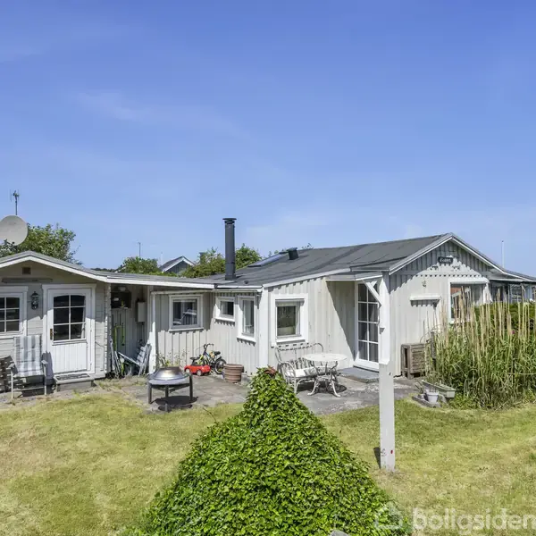 Et lille hvidt sommerhus med terrasse står på en velholdt græsplæne. Der er havemøbler foran huset, og himlen er klar.