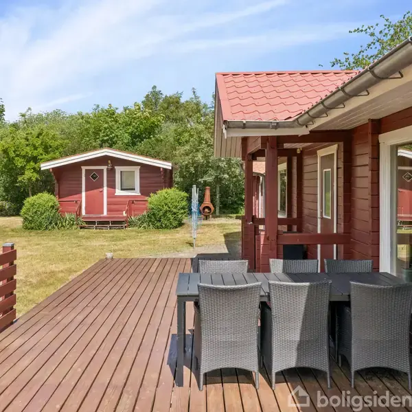 Rødt sommerhus med træterrasse. På terrassen står et sort havemøbelsæt. Foran huset ses en græsplæne med et mindre rødt skur. Omgivelserne består af grønne buske og træer.