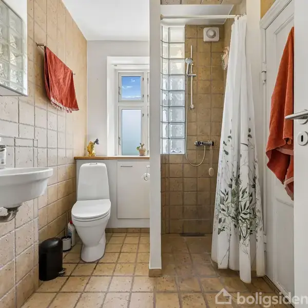 Badeværelse med en hvid håndvask og toilet. Brusekabine til højre med et forhæng. Beige fliser på vægge og gulv. Et vindue bagtil giver lysindfald.