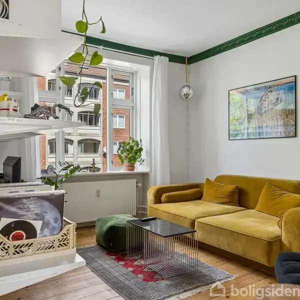 En gul sofa står foran en væg med en plakat. Ved siden af er et vindue med planter. Et lille bord og tæppe findes på gulvet. Hylder hænger på væggen.