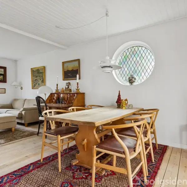 Et spisebord af træ står i et lyst rum med seks stole omkring. I baggrunden er en lys sofa, lamper, vægdekorationer og et rundt vindue med gitter. Gulvtæppe under bordet.