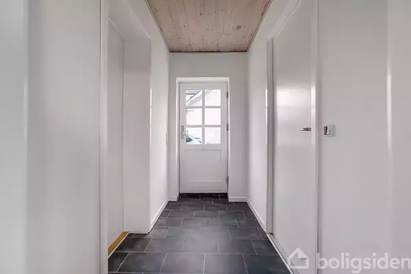 En hvidmalet gang med mørkt flisegulv og trægulvloft fører til en glasdør i et hjemligt miljø.