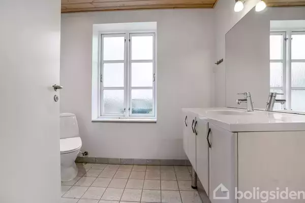 Toilet står ved siden af en dobbelt vask i et lyst badeværelse med flisegulv. Et vindue i midten giver naturligt lys.