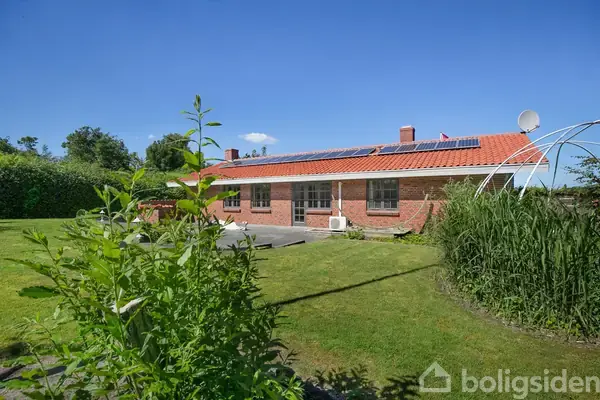 Et rødt murstenshus med rødt tegltag ligger i en have omkranset af tætte, grønne buske. En flisebelagt indkørsel fører op til en garage ved siden af huset.