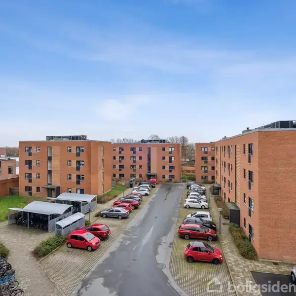 Røde murstensbygninger står rundt en parkeringsplads fyldt med biler i forskellige farver. Området er en boligkvarter med græsplæner og cykelstativer, set under en klar, blå himmel.