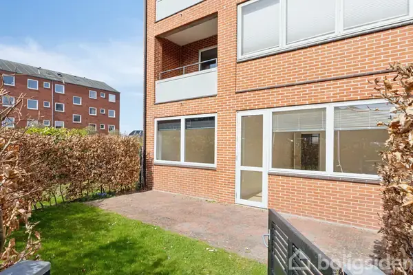 Rødstensbygning med store vinduer og en dør åbner ud til en flisebelagt terrasse. Omgivet af grøn have og hæk. I baggrunden ses en anden boligblok.