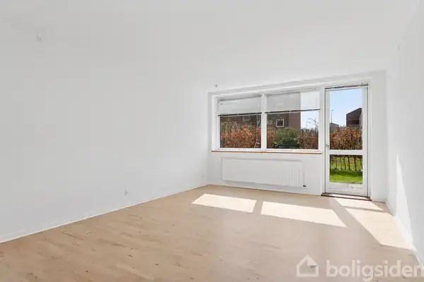 Et lyst, tomt rum i en bolig med trægulve, store vinduer og dør ud til en have. Sollys strømmer ind gennem vinduerne, og udenfor ses grønne buske.