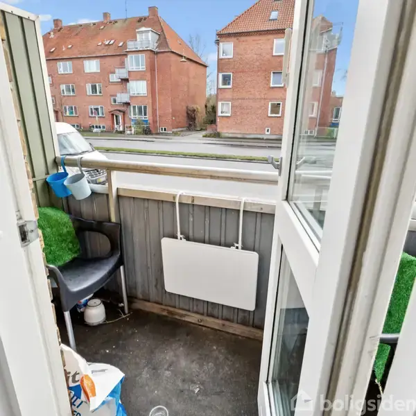 En balkon med åben dør fører ud til en lille udendørs plads med stole. Udenfor ses en rolig vej og murstensbygninger. Græsbeklædning findes på gelænderet.