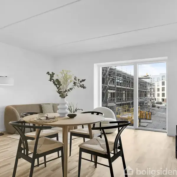 En indrettet stue har et rundt spisebord med stole, en sofa og en lampe. Et vindue viser en byggeplads udenfor. Der er ingen tekst i billedet.