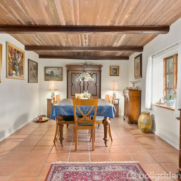 Et spisebord dækket med en blå dug står i centrum af en spisestue. Stole omgiver bordet, og rummet er udsmykket med kunstværker, træmøbler og lamper. Vinduet giver naturligt lys.