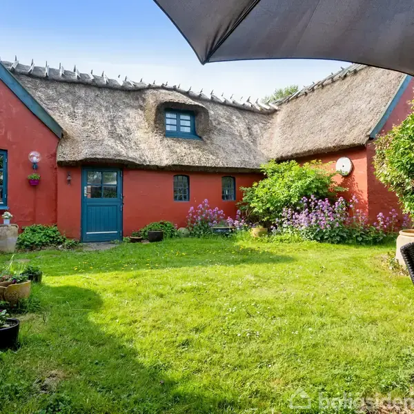 Rødt bindingsværkshus med stråtag står i en have med græsplæne, blomsterbede og krukker. I forgrunden ses havemøbler og en parasol, der skaber en hyggelig stemning.
