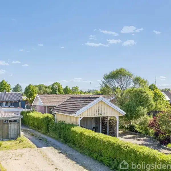 En carport står ved siden af en grusvej, omgivet af grønne hække og træer. I baggrunden ses et beboelseshus, yderligere huse og grønne områder under en klar blå himmel.