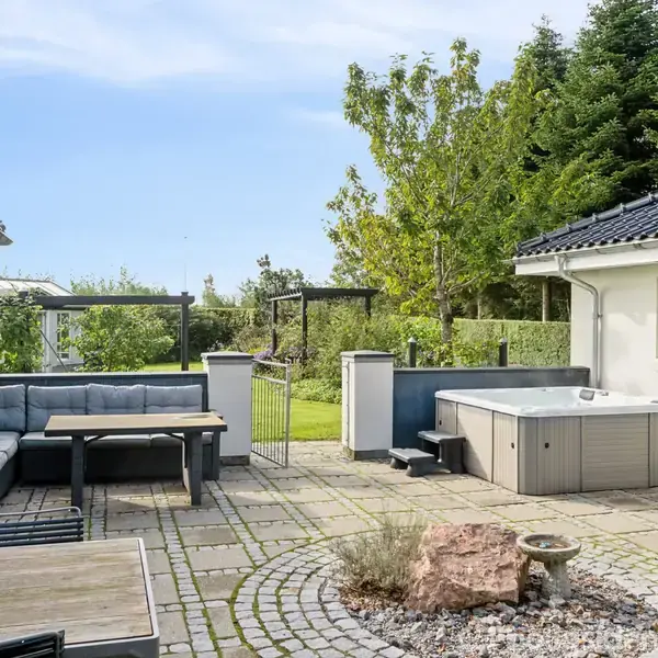 Terrassen har et boblebad og sofaarrangement. Den er omgivet af to huse og en have med træer, hvilket skaber en afslappende atmosfære.
