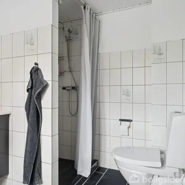 Toilet er installeret ved siden af en bruser med hvidt forhæng; hvide fliser dækker væggene, mens sorte fliser dækker gulvet; et spejl og vaskeskab er ved siden af.