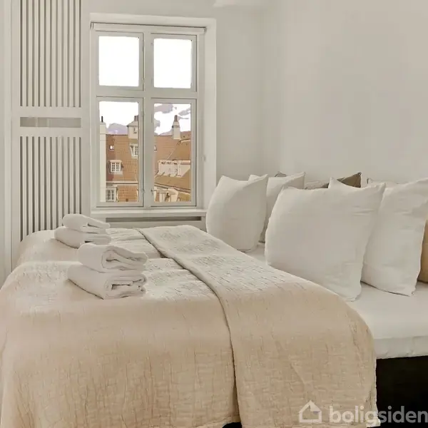 Sengen er redt med hvidt sengetøj og beige tæpper, to håndklæder ovenpå. Vinduet viser hustage udenfor. Rumindretningen er lys og minimalistisk.