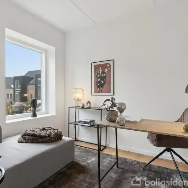 Et lyst arbejdsrum med et skrivebord med en lampe og dekorationer, en stol ved siden af en bænk under et vindue. Vægdekorationer og bogreol supplerer rummet.