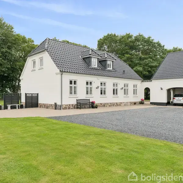 Hvidt hus med mørkt tag står ved siden af en garage med to biler. Græsplæne og træer omgiver bygningerne i et åbent, naturskønt område.