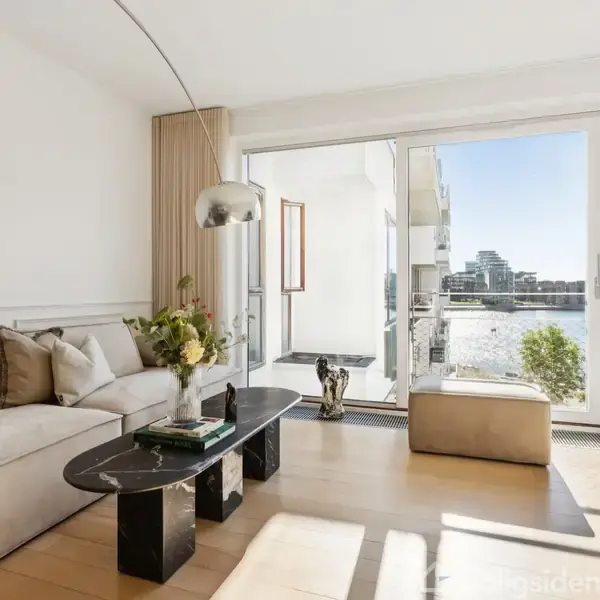 Stue med beige sofa og sort sofabord, sollys skinner ind gennem et stort vindue med udsigt til en balkon og vand. En moderne lampe hænger over sofabordet.
