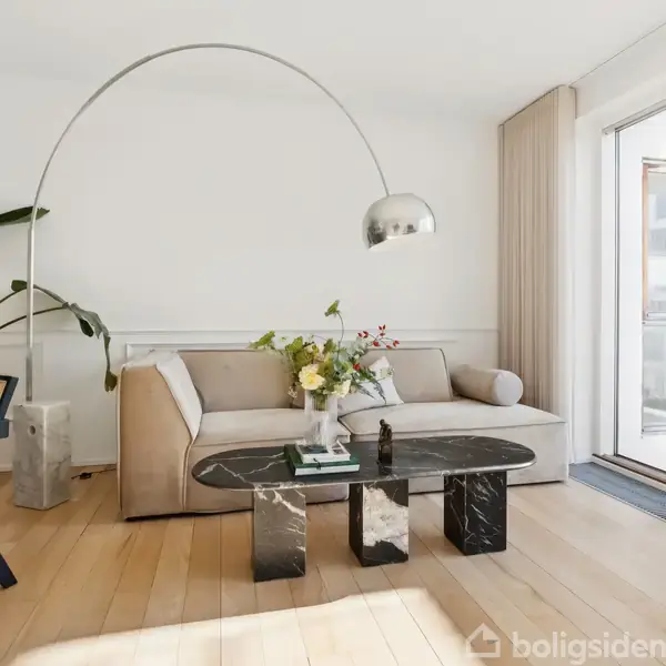 En beige sofa står i en moderne stue med en buet gulvlampe over sig. Et sofabord med blomster og bøger står foran, og der er store vinduer med udsigt til en altan.