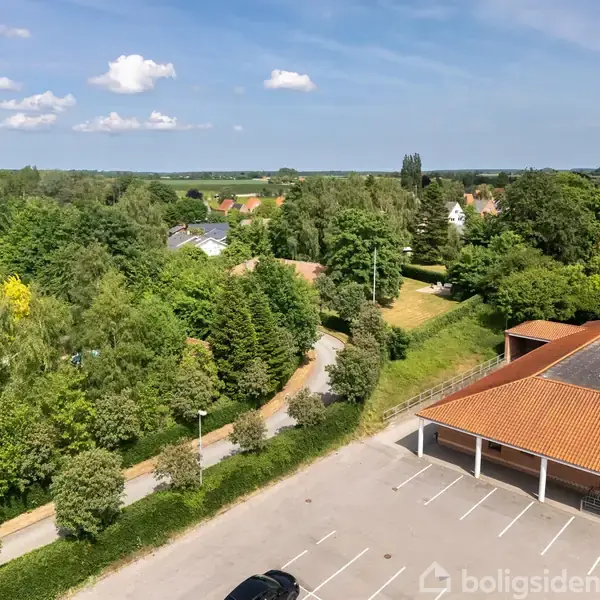 Bygning med rødt tag grænser op til parkeringsplads, omgivet af frodige grønne træer og en vej. I baggrunden ses et beboelsesområde og en blå himmel med spredte skyer.