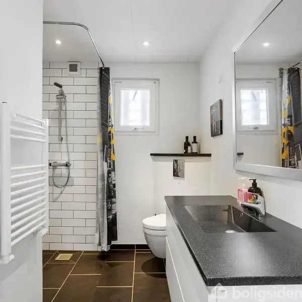 Badeværelse med sort bordplade-vask, spejl, og brusekabine med hvid flisevæg. Bruser og håndklæder ses ved siden af et lille vindue. Brun flisegulv og radiator ved døren.