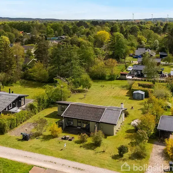 Et sommerhus ligger blandt andre huse i et frodigt, grønt område med træer og buske. En grusvej løber gennem området, og en blå bil er parkeret nær et hus.