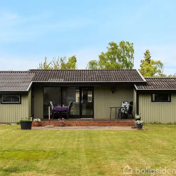 Et enkelt, étplans hus med gråt tag står stille i en frodig have. En lille terrasse foran huset har en spiseplads, og omgivelserne er præget af grønne træer og buske.